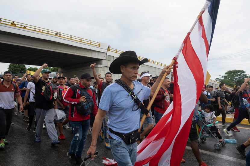 Un migrante lleva una bandera estadounidense mientras arrastra equipaje durante una caravana...