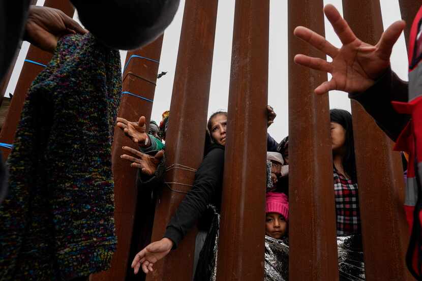 Migrantes extienden los brazos a través de una valla fronteriza para tomar prendas de ropa...