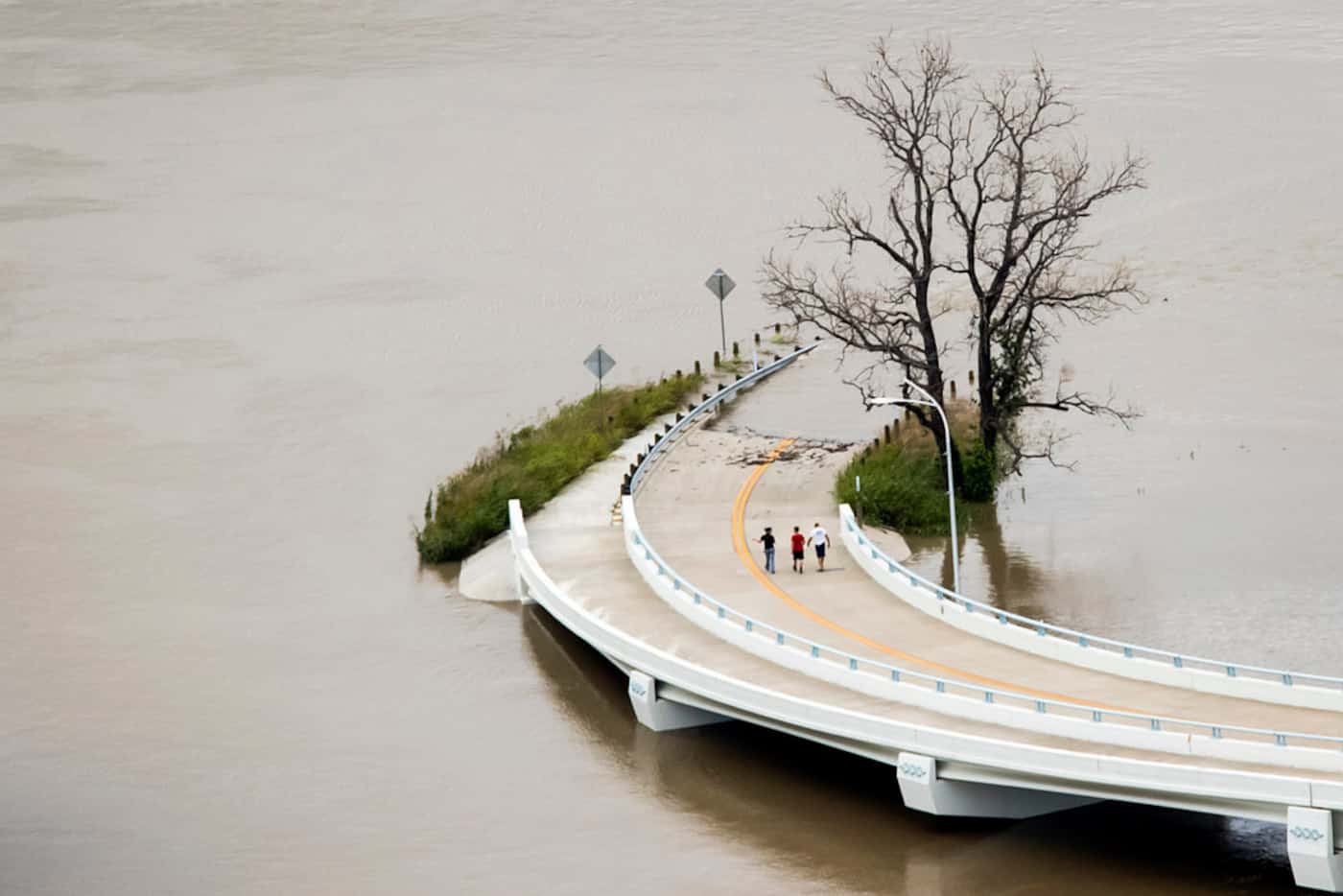 Floodwaters from the swollen Trinity River surround a roadway as people walk from the Sylvan...
