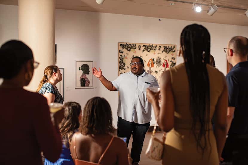 Man standing in art gallery motions to artworks as people look on.