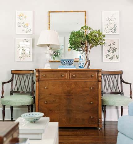 Wooden chest flanked by two antique looking chairs and topped with a lamp, flowers, and more...