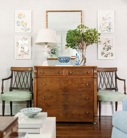 Wooden chest flanked by two antique looking chairs and topped with a lamp, flowers, and more...