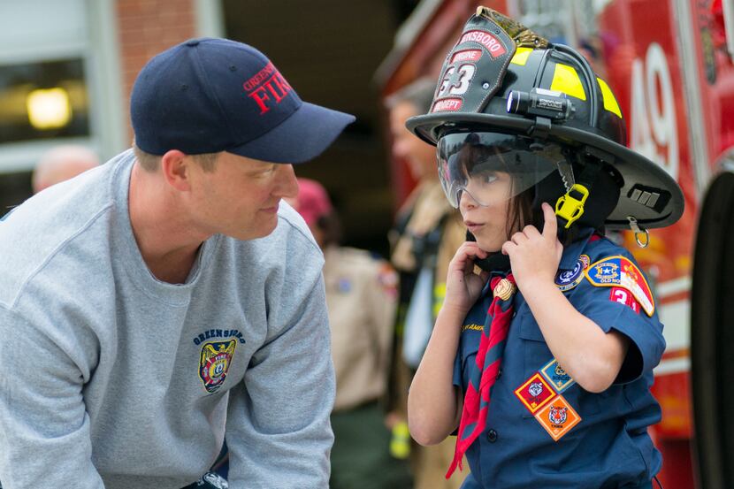 A young scout in uniform speaks with a firefighter and tries on his helmet.