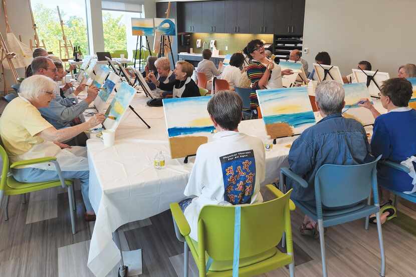 Senior citizens sit at tables painting a seascape.