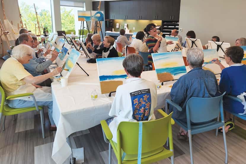 Senior citizens sit at tables painting a seascape.
