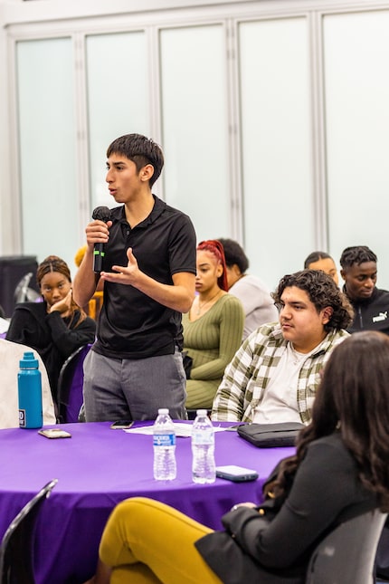 A young male college student holds a microphone and talks to a group at a round table.