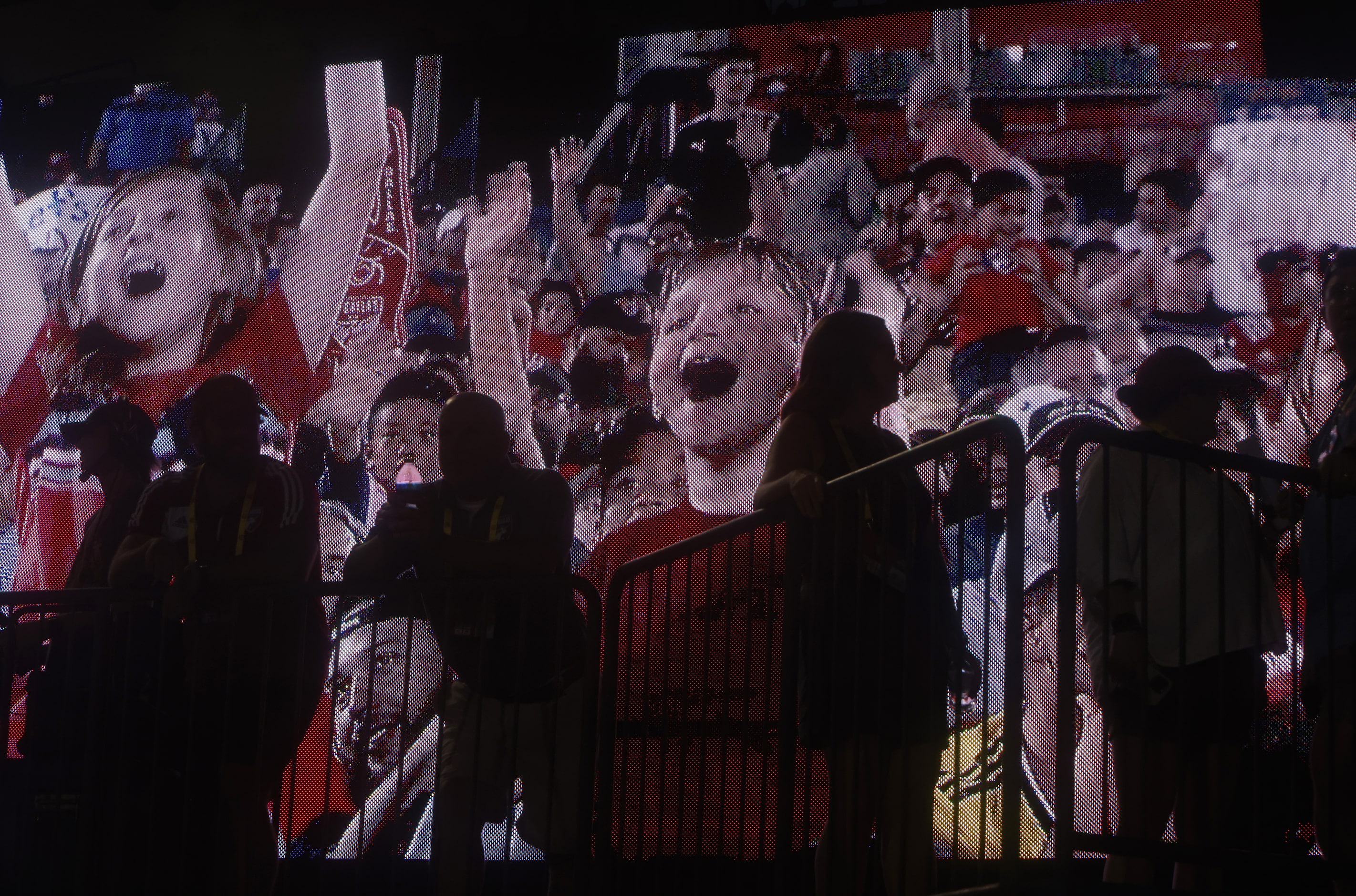 Young FC Dallas fans are projected on a large projection screen behind fans standing along a...