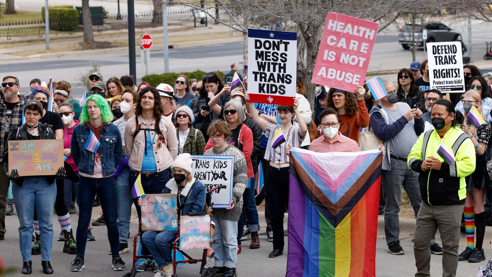 Attendees hold signs and carry flags as they listen to a speaker during the "Trans Kids Cry...