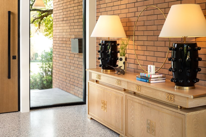 Buffet in light wood against a brick wall, with two black lamps and a stack of books, next...