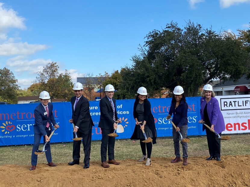 Six people in hard hats and suits hold shovels in front of dug up ground to celebrate...