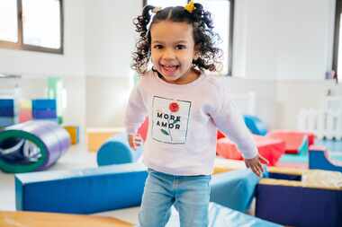 A cute little girl is smiling and jumping on soft blocks in a preschool classroom.