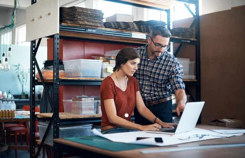 A man and a woman look at a laptop together, standing in front of a shelf with tools and...