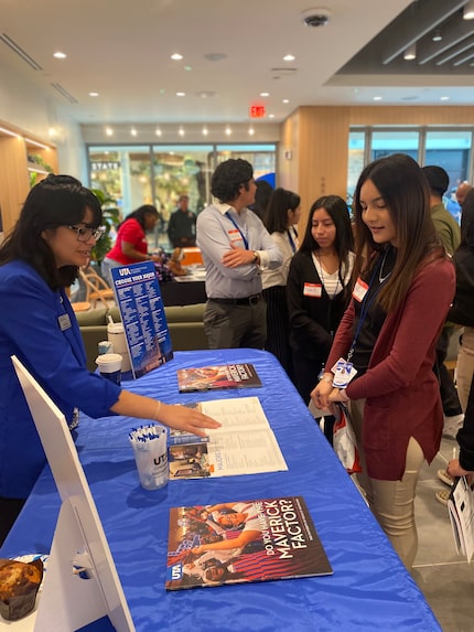 Student takes a flyer from a long table covered in blue tablecloth during college readiness...