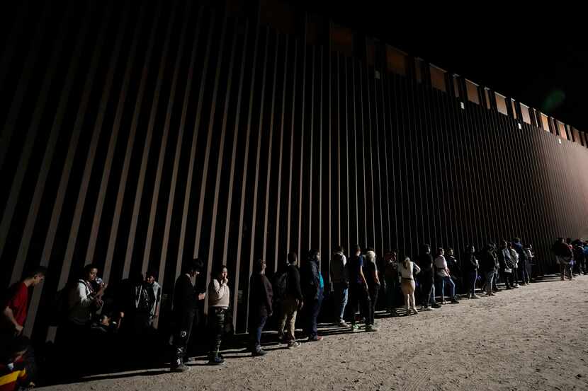 Un grupo de personas forman una fila junto a un muro fronterizo mientras esperan para...