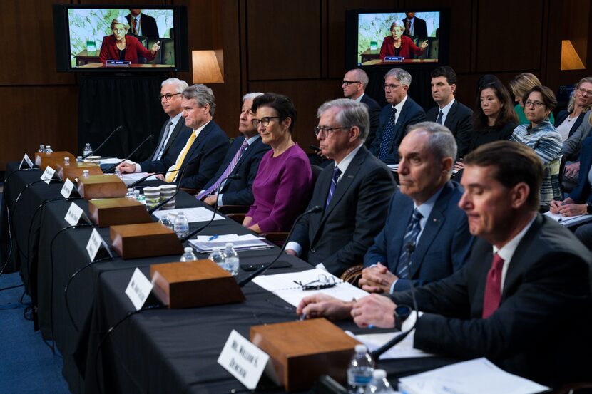 Sen. Elizabeth Warren, D-Mass., on a screen in the background, questions witnesses about...