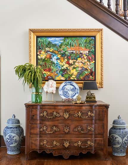 A brightly colored painting of a waterfall hangs over an antique credenza, two blue and...