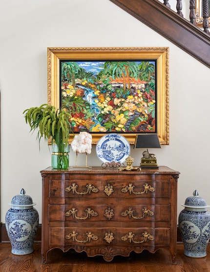 A brightly colored painting of a waterfall hangs over an antique credenza, two blue and...