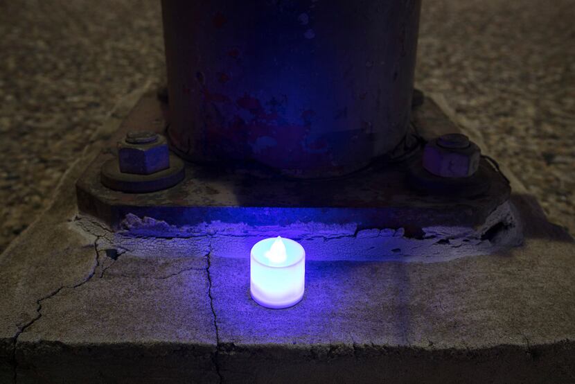 At 9:43 p.m., on a quiet sidewalk, a single blue electric candle glows next to a light pole...