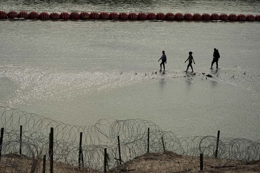 Migrants walk past large buoys being used as a floating border barrier on the Rio Grande,...
