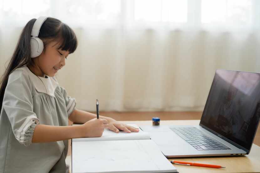 Young Asian girl wears white headphones while working on homework at a laptop.