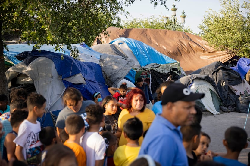 Tents and tarps behind children who are lining up to tell a woman their names and what toys...
