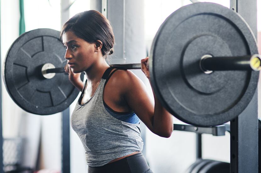 A woman lifts a barbell in a gym while wearing a gray tank top.