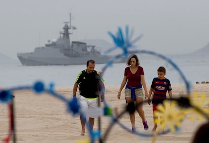 A family walks on the shore of Copacabana beach backdropped by a Brazilian navy vessel, in...