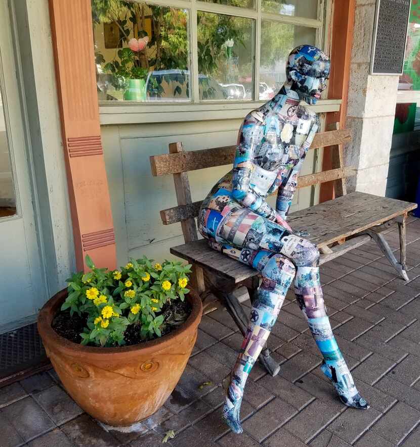 A decoupaged sculpture of a woman on a bench sits outside the office of the twice-weekly...