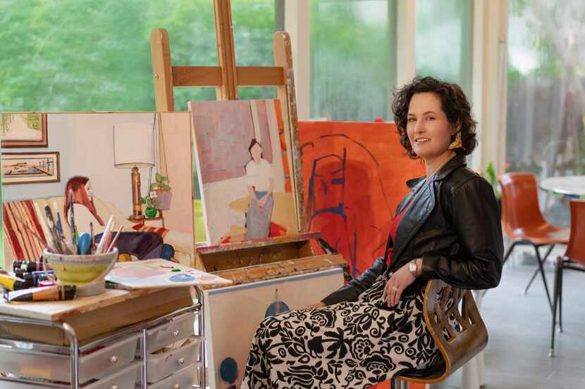 Liz Jordan sitting in a chair surrounded by canvases and art supplies, before a glass window...