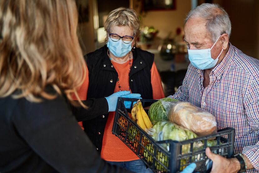 A neighbor delivers a box of groceries to a senior couple wearing face masks during the...