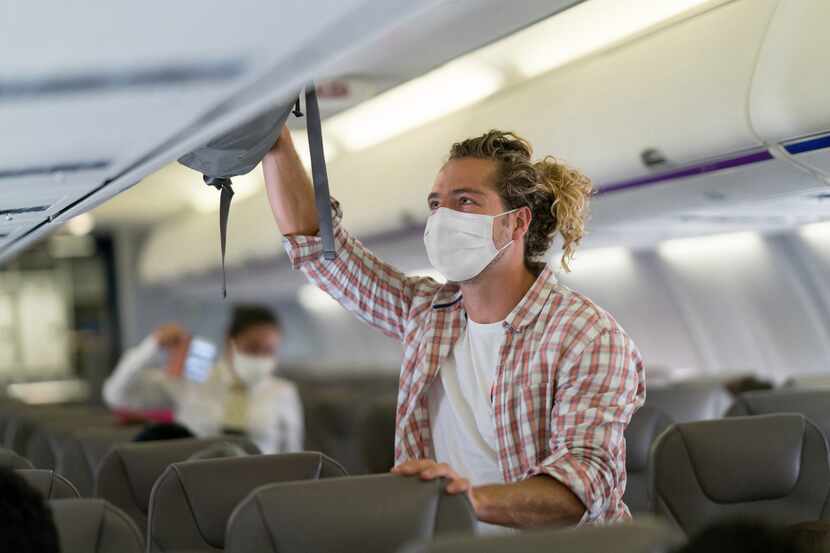 Un pasajero con mascarillas ubica su equipaje en el compartimento de la cabina de un avión,...