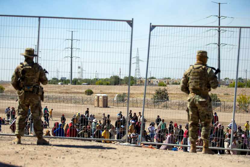 FILE - Migrants wait in line adjacent to the border fence under the watch of the Texas...