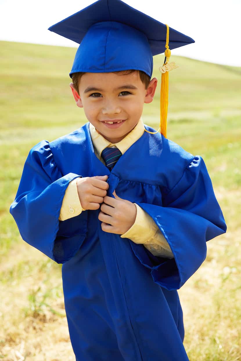 A Hispanic kindergartener stands in a field wearing his bright blue graduation cap and gown.