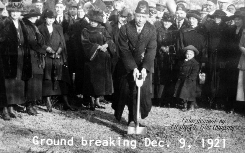 In a black and white image from 1921, a man digs a shovel into the ground with a crowd...