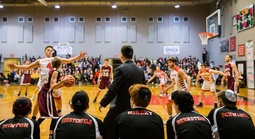 Yavneh basketball players wearing yamakas watch from the bench as their teammates play in...