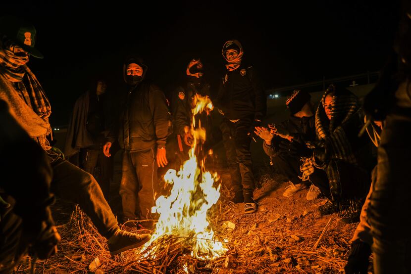A group of migrants warm themselves with burning trash on the banks of the Rio Grande river...