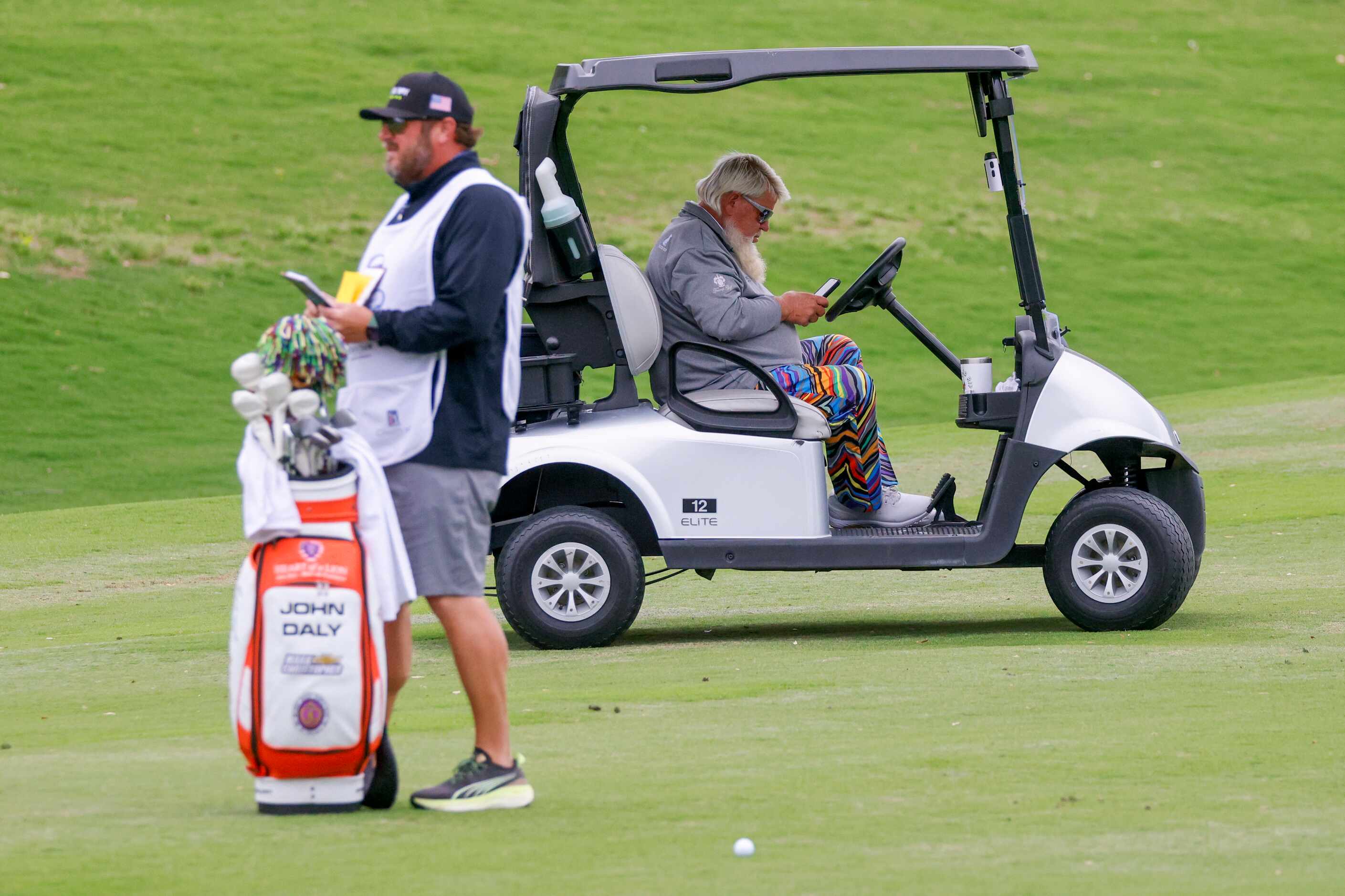 John Daly looks at his phone as his caddie checks a yardage book on the 18th fairway during...