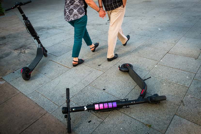 A couple walks past a pair of rental scooters littering Commerce Street in downtown Dallas...