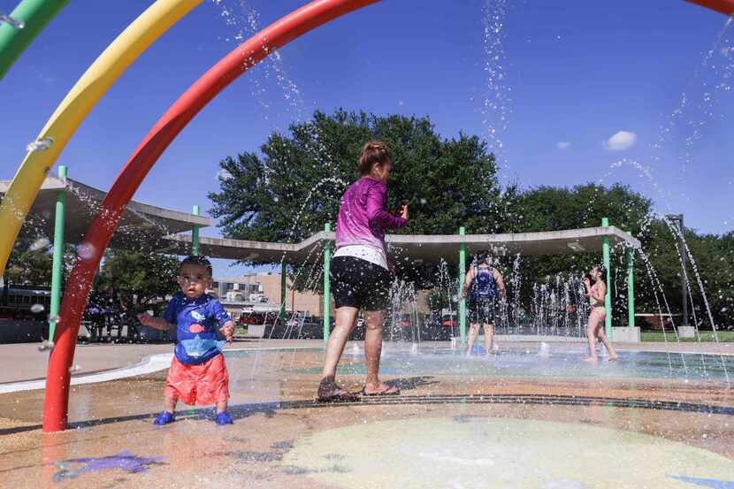 Adults and kids cool off from heat in a splash pad at Ferguson Park while a clear sky over...