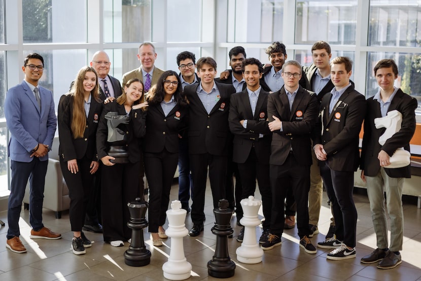 The UT Dallas chess team stands with their coaches in front of large chess piece decorations...