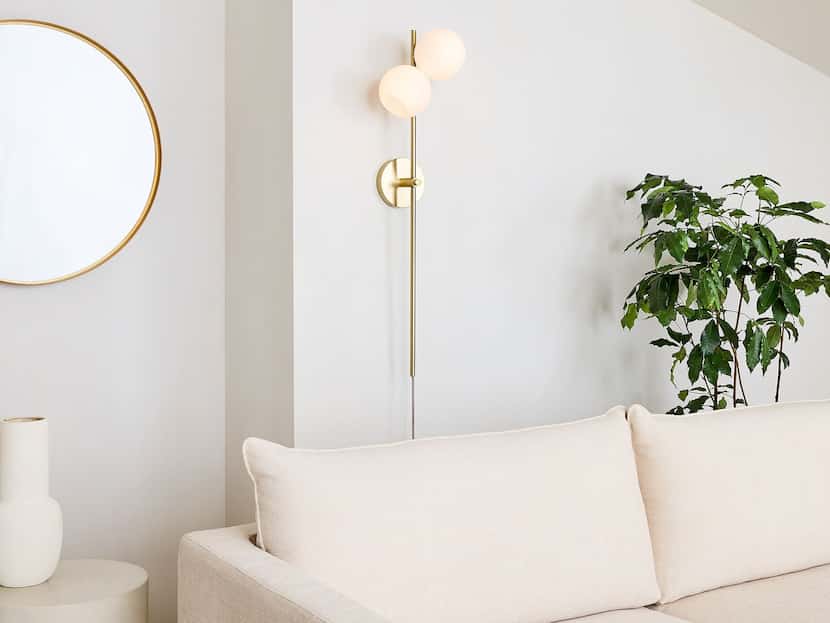 White wall with two-bulb gold sconce, white sofa, plant, gold mirror and white vase