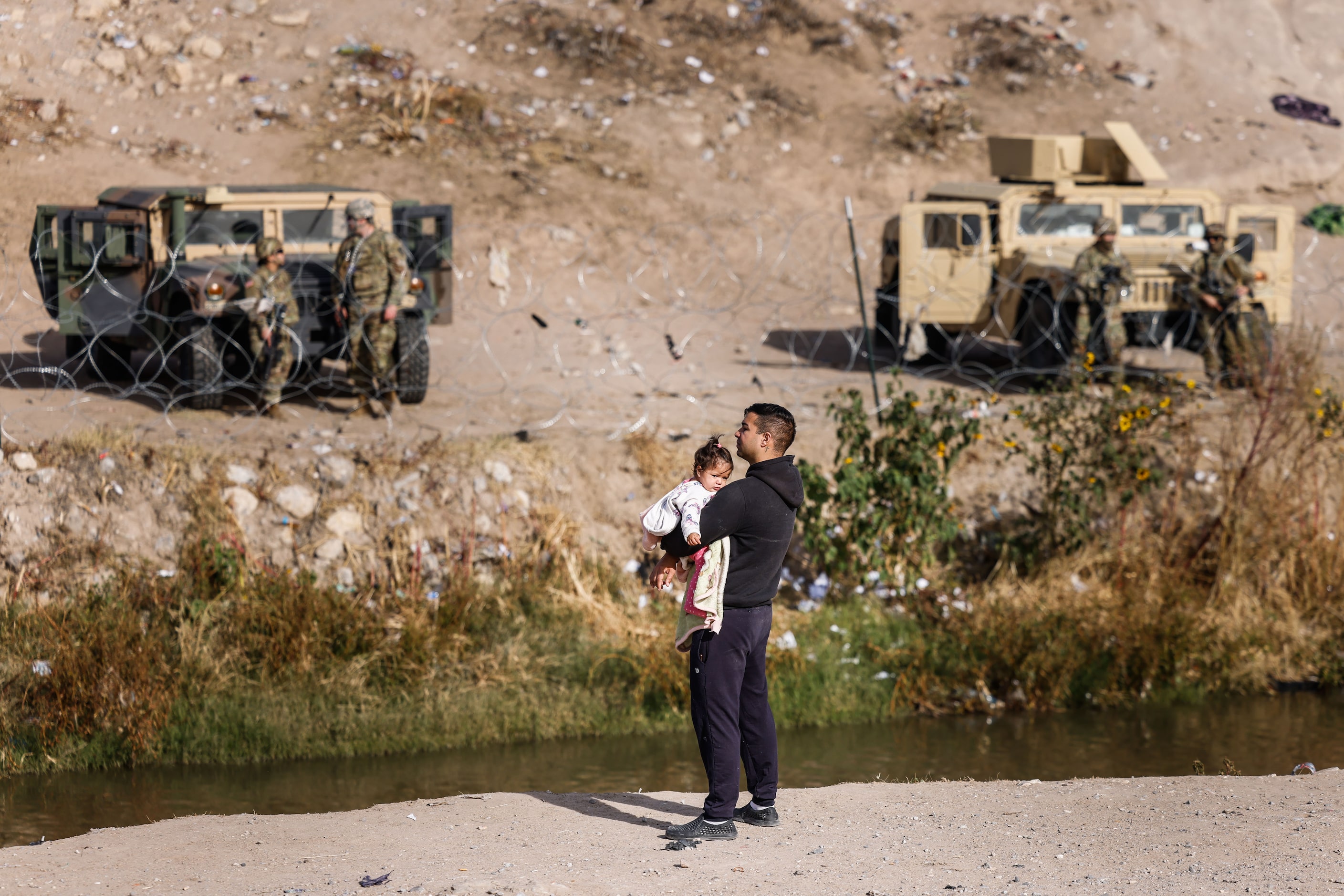 A migrant with a baby in his arms watches the scene on the other side of the border in El...