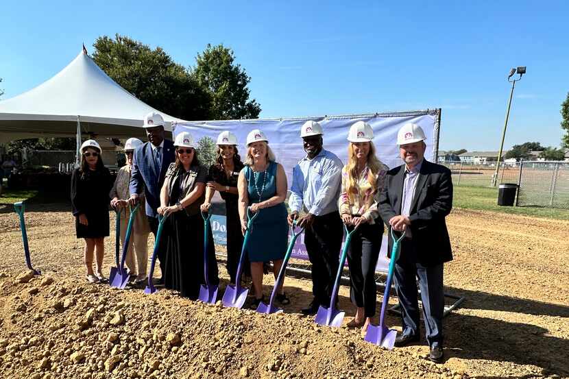 Nine people in hard hats and purple shovels pose on a dirt field.