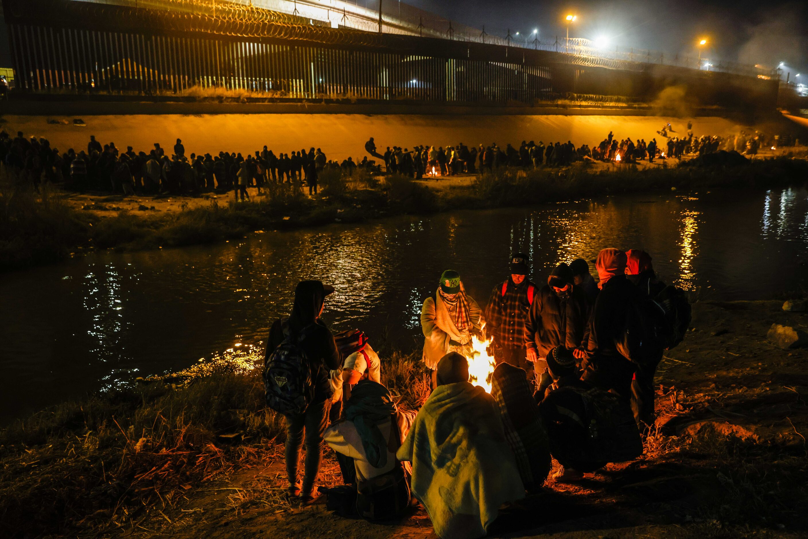 A group of migrants warm themselves with burning trash on the banks of the Rio Grande river...