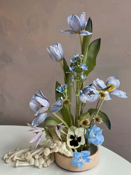 Blue floral arrangement in a shallow tan dish with a collection of mushrooms spilling out of...