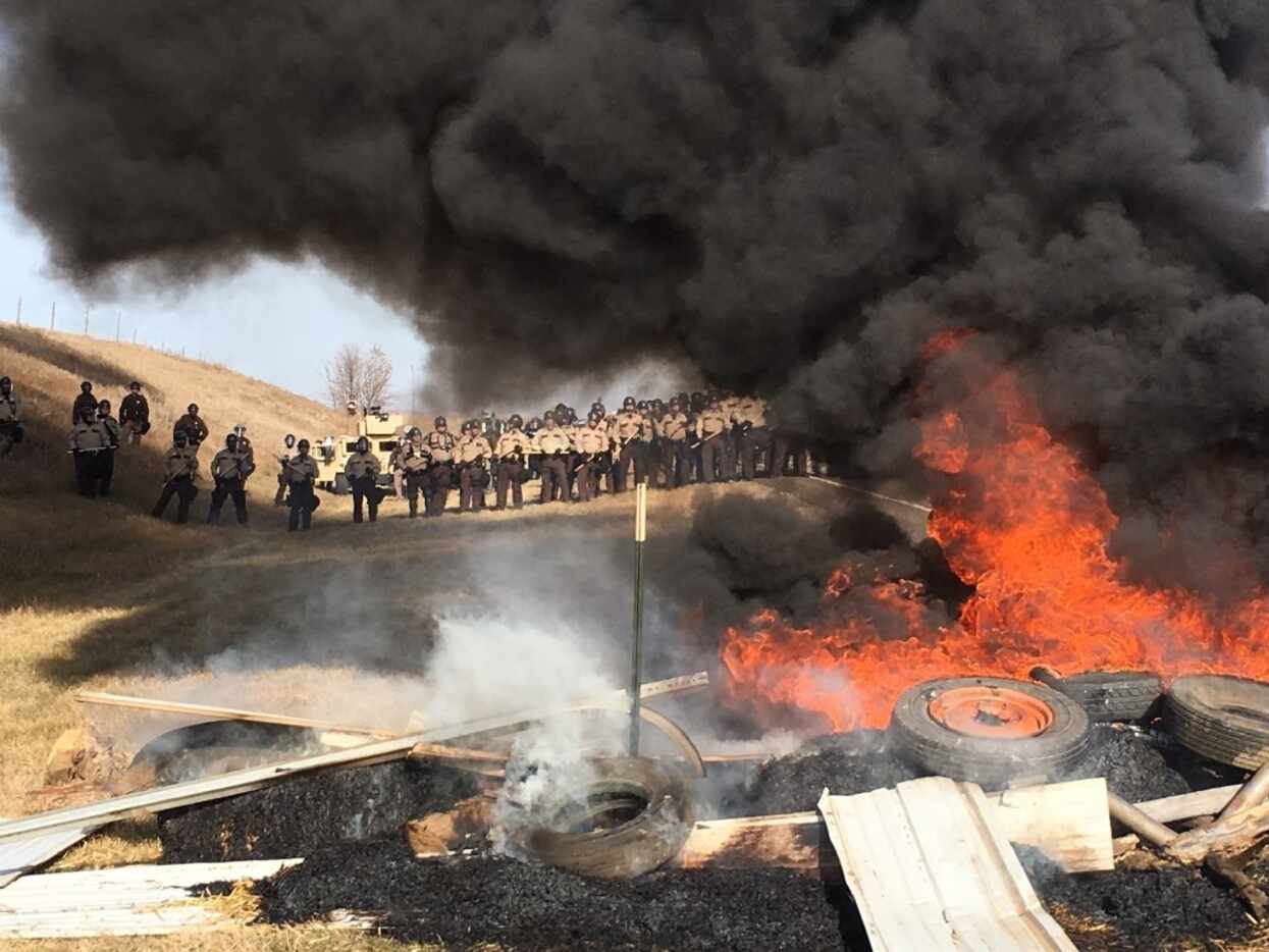 Tires burn as armed soldiers and law enforcement officers stand in formation to force Dakota...