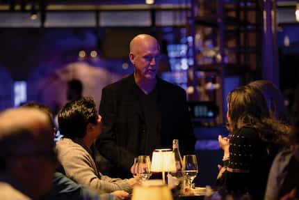 Man dressed in black greets a couple dining at a table in a dimly lit venue.