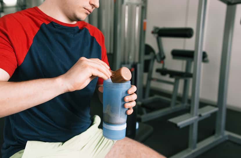 A man pours protein into a shaker after a workout while sitting on a bench in the gym.