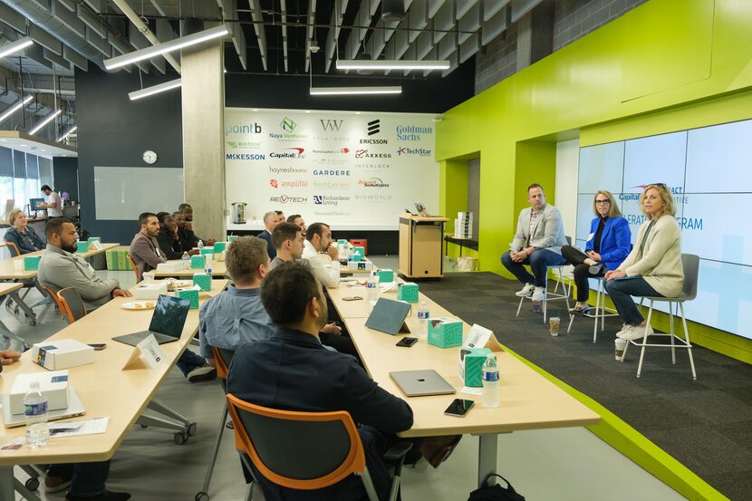 Three people sit on stools in front of people sitting at long tables in a conference room.