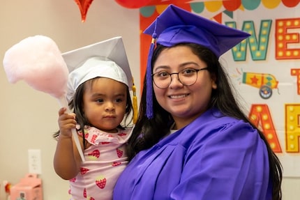 Young mother in a blue graduation cap and gown holds her infant, who is wearing a white...